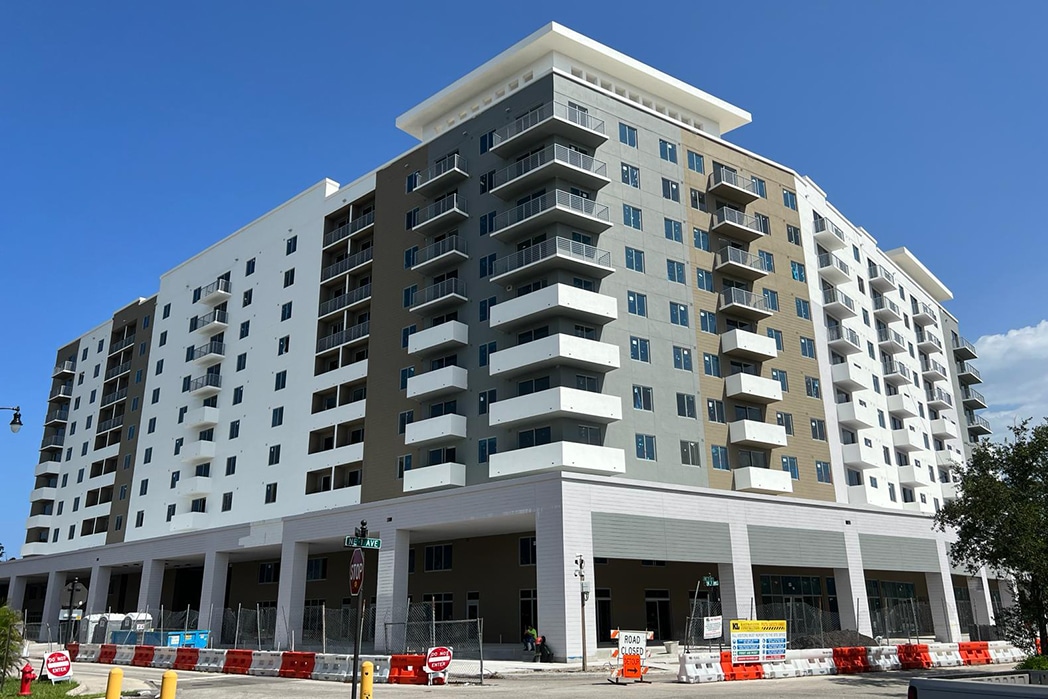 A modern mid-rise apartment building under construction with multiple balconies, large windows, and a sleek facade. Construction barriers are set up along the street.
